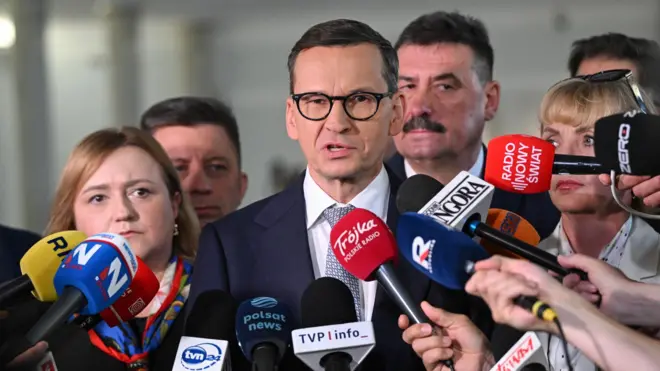 Mężczyzna z ciemnymi, krótkimi włosami ,w czarnych okularach, białej koszuli i w niebieskiej marynarce stoi w otoczeniu pięciu osób, przed nimi znajdują się mikrofony prasy
