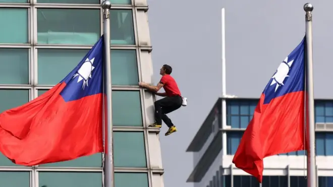 Alex Honnold, wearing a red T-shirt and black trousers, climbs a skyscraper. Two large flags of Taiwan flutter in front of the building.