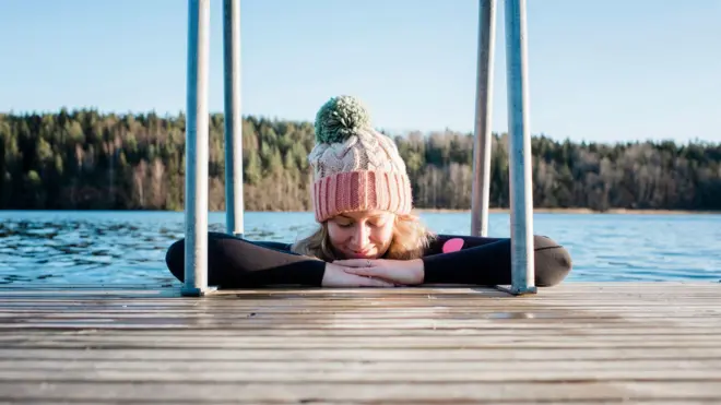 Kobieta w czapce opiera się na molo, ma zamknięte oczy, w tle widać wodę i las