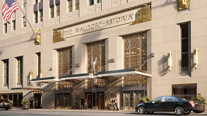 Fachada del Waldorf Astoria en Nueva York, de color arena con elementos dorados, y la bandera de Estados Unidos colgada de un mástil al costado del letrero donde se lee el nombre del hotel.