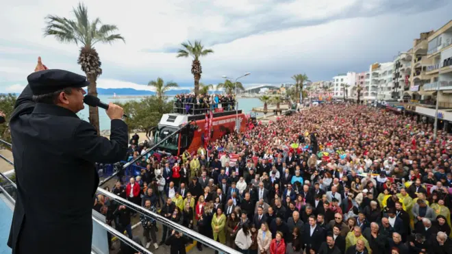 Özgür Özel kasketli bir halde otobüsün üzerinde elinde mikrofonla kalabalık bir kitleye konuşuyor