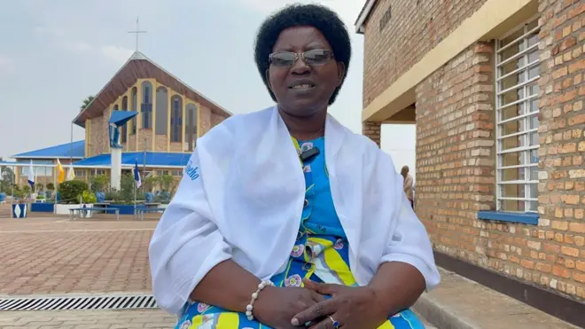 Nathalie Mukamazimpaka yicaye yiteye fulari y'umweru, yambaye ishapule ku kuboko, inyuma ye hari Kiliziya iriho umusaraba. 