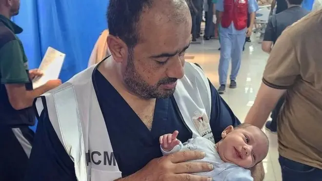 Dr Muhammad Abu Salamiya holding a baby in a bustling hospital ward