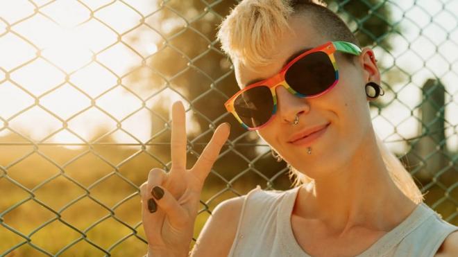 Pessoa branca e loira com parte dos cabelos raspados,jogo de máquina caça níquel de moedaóculos escuros e piercings no septo e na boca,jogo de máquina caça níquel de moedafrente a uma gradejogo de máquina caça níquel de moedaum dia ensoladarado