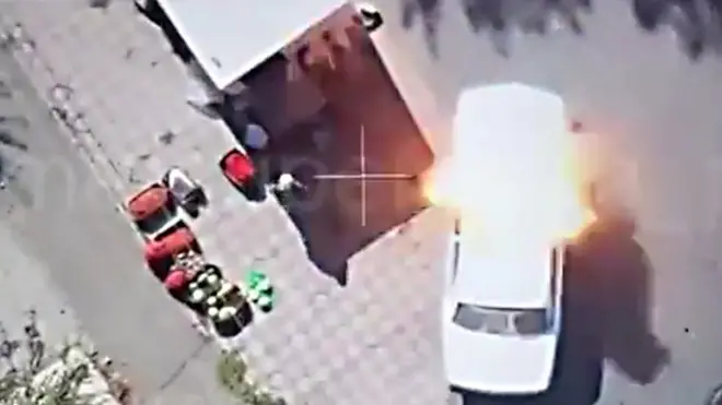 A screengrab of drone footage showing crosshair sights and an overhead view of white car on a street at the moment when an explosive detonates on its roof