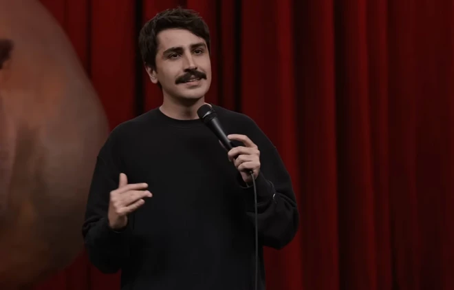 Deniz Göktaş'ın Harbiye'de sahnelediği gösteri sırasında kırmızı bir perde önünde konuşuyor. Bıyıklı ama genç bir görüntüsü var. Bir elinde mikrofon tutuyor.