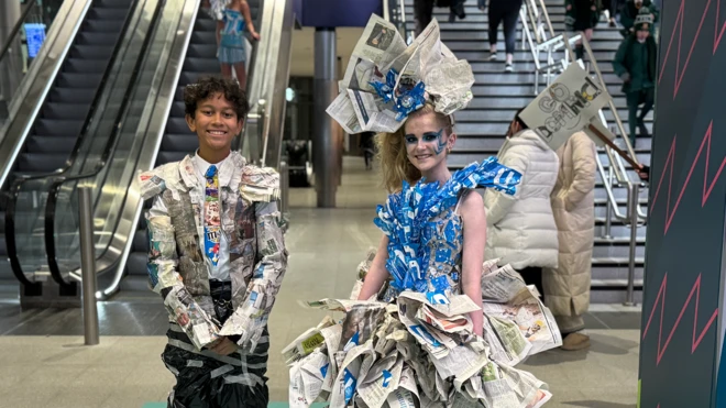 Dominic and Aoife are wearing sustainable outfits they made, with an assortment of newspaper clippings, plastic bags, train tickets and tape. They are standing and smiling at the camera. 