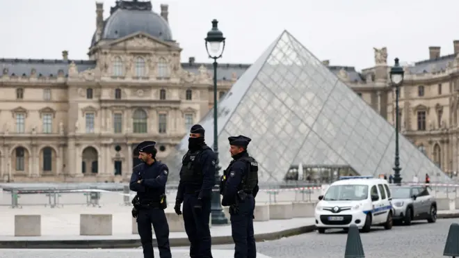 Три французских полицейских в синей форме на фоне здания Лувра и стеклянной пирамида И.М. Пэя, также видна полицейская лента