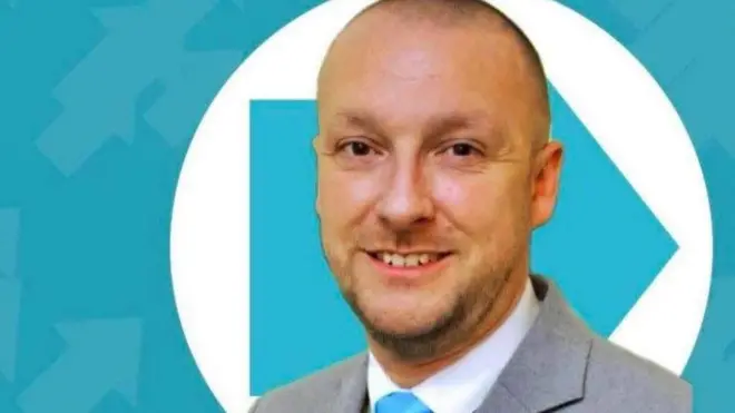 Стівен Мусделл, якому за 30, з поголеною головою та щетиною, посміхається в камеру в сірому костюмі зі світло-блакитною краваткою. На задньому плані - логотип Reform