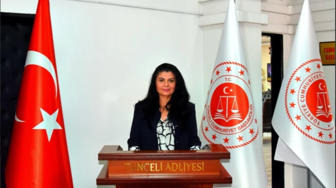 Uzun siyah saçlı, siyah takım elbiseli bir kadın kürsüden konuşma yapıyor. Sağ yanında Türkiye bayrağı var. 