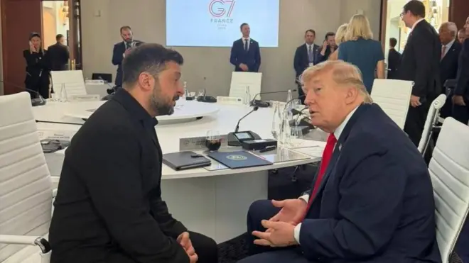 Зеленський у чорному костюмі та Трамп сидять поруч за круглим столом і ведуть розмову, нахилившись одне до одного. На столі видно документи, планшети, мікрофони, келихи та пляшки з водою. На задньому плані — інші учасники саміту, які стоять і спілкуються, а також екран із написом G7 France.