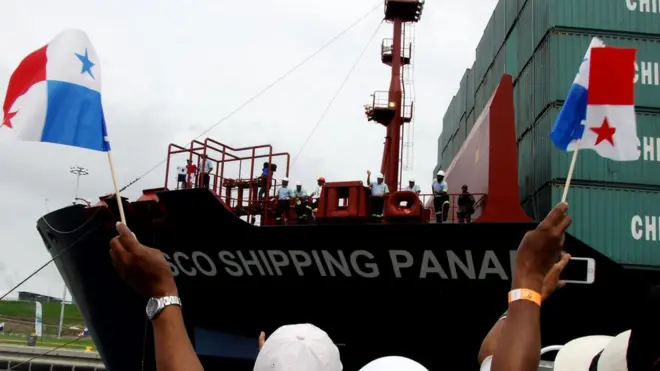 Varias personas sostienen banderas frente a un buque atracado en el canal de Panamá.