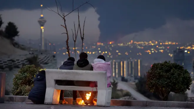 Three Tehran residents sit on a bench overlooking smoke billowing in the distance at night on 8 March.