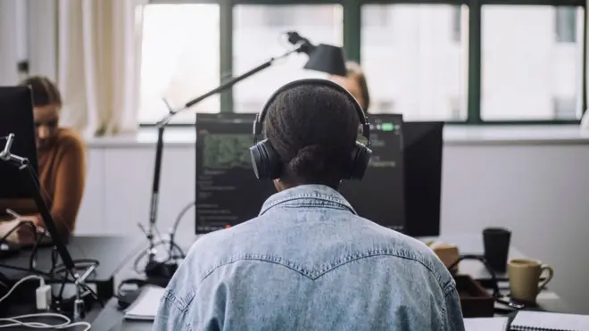 一位穿著牛仔外套的女子從背後被拍下，正戴著耳機在電腦前工作。