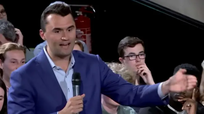 查理·柯克穿著藍色西裝襯衫，手拿麥克風，一頭深棕色短髮，正對著麥克風做著手勢、說話。他身後坐著一些人，正在觀看。