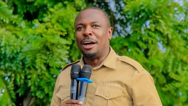 John Heche afashe indangururamajwi ebyiri munsi y'umunwa we. Yambaye ishati y'ibara ry'ikijuju yanditseho izina rye.