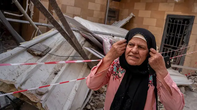 امرأة تقف أمام مبنى متضرر، وخلفها ركام وألواح معدنية منهارة وشريط تحذيري أحمر وأبيض، بعد غارة إسرائيلية في صور بجنوب لبنان. ترتدي المرأة غطاء رأس أسود وقميصاً وردياً مطرزاً، وتمسك هاتفاً بيدها بينما تضع يدها الأخرى قرب أذنها.