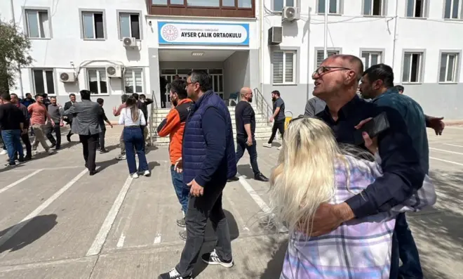 Saldırının yaşandığı okulun önünde toplanan aileler birbirlerine sarılarak destek veriyor. Arkada okulun ismi var.