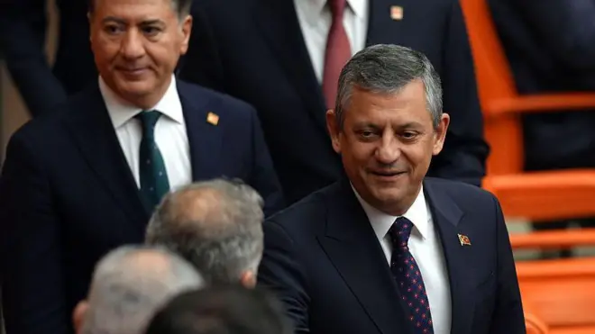 Özgür Özel bir siyasetçinin elini sıkıyor. Burası meclis. Arkada turuncu deri koltuklar var.
