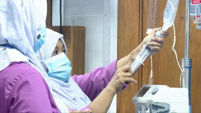 মাস্ক পরা একজন নার্স স্যালাইনে ইনজেকশন পুশ করছেন। পাশে দাঁড়িয়ে আছেন আরেকজন নার্স।