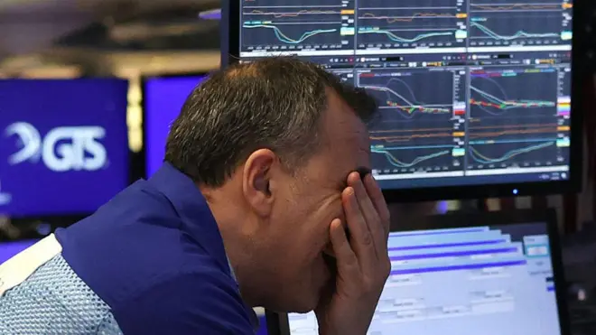 A trader covering his face with his hand. He is wearing a blue shirt and is leaning on a desk. In the background four laptops with graphics on the screen. 
