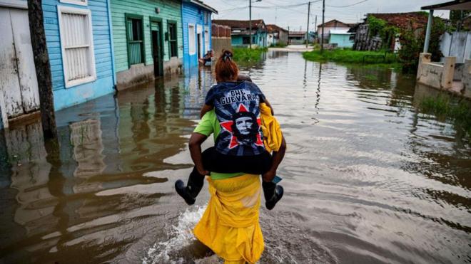 Un hombre carga a una mujer en su espalda mientras camina por el agua en un área inundada de Batabanó, provincia de Mayabeque, Cuba, el 28 de agosto de 2023, mientras la tormenta tropical Idalia se acerca al extremo occidental de la nación insular.