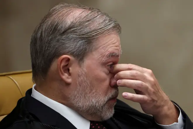 Toffoli sentado durante sessão do STF, fotografado de perfil, leva a mão esquerda ao nariz e aos olhos