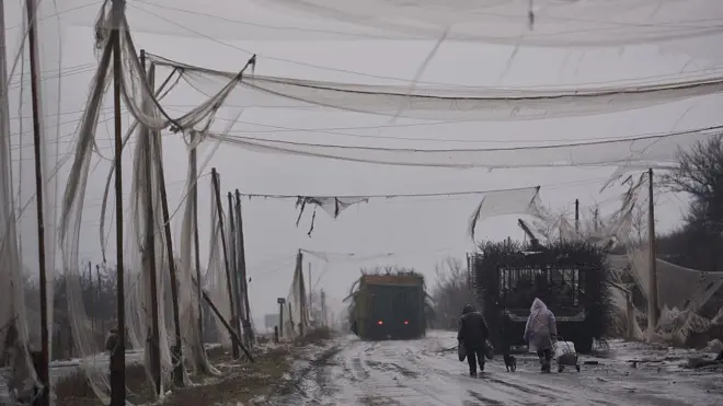Мокра і брудна дорога з військовою технікою, накрита пошматованими білими сітками. Дорогою, повз танк із протидроновим "мангалом", йдуть двоє цивільних із візком і собакою.