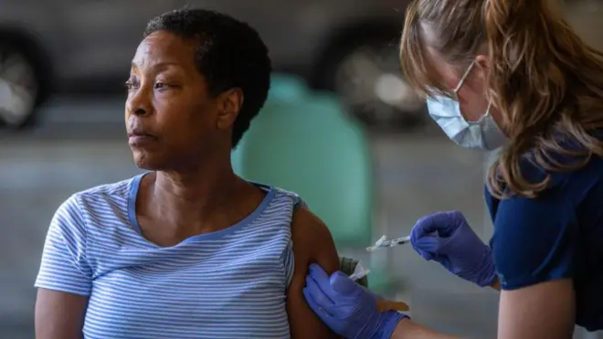 Une femme vêtue d'un t-shirt rayé bleu et blanc est assise et détourne le regard pendant qu'un professionnel de santé lui administre un vaccin.