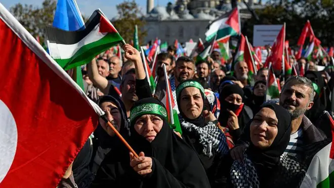 Filistin destekçileri ellerinde Türk ve Filistin bayraklarıyla mitinge katılıyor. Önde Başörtülü kadınlar var.