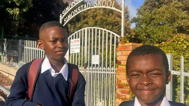 Les frères Anele (à gauche), qui porte un sac à dos, et Lifalethu Mbasana, vêtus de leur uniforme composé d'un pull bleu marine et d'une chemise blanche, devant le portail de leur école à Simon's Town.