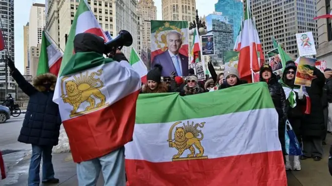 Ellerinde ve üzerlerinde aslan ve güneş motifli İran bayrakları taşıyan muhalif göstericiler eylem yapıyor. Bir tanesinin elinde megafon var ve kalabalığa seslendiği görülüyor.