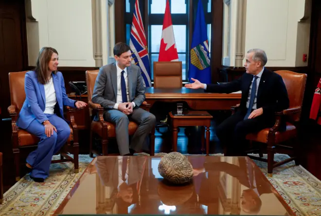 Смит, Эби и Карни сидят на креслах спиной к длинному столу, Смит и Эби с одной стороны (на фото слева), Карни с другой, на некотором расстоянии от них. За столом стоят флаги, за ними окно. На переднем плане еще один стол, в котором отражаются политики, на нем лежит сувенир в виде камня, на котором вырезаны извилистые линии.