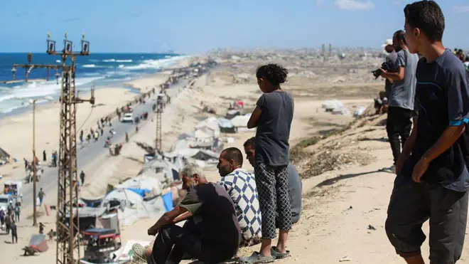 يشاهد فلسطينيون أشخاصاً يصلون مع ممتلكاتهم على طريق ساحلي شمال غرب مخيم النصيرات للاجئين، أثناء نزوحهم جنوباً من وادي غزة.