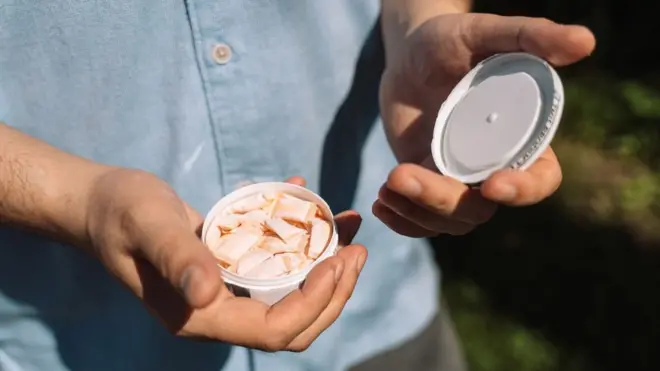 ਇੱਕ ਆਦਮੀ ਕੈਮਰੇ ਵੱਲ ਨਿਕੋਟੀਨ ਪਾਊਚਾਂ ਦਾ ਇੱਕ ਛੋਟਾ ਜਿਹਾ ਡੱਬਾ ਦਿਖਾ ਰਿਹਾ ਹੈ, ਜੋ ਦੇਖਣ ਵਿੱਚ ਬਹੁਤ ਛੋਟੇ ਸਿਰਹਾਣਿਆਂ ਵਰਗੇ ਲੱਗਦੇ ਹਨ।