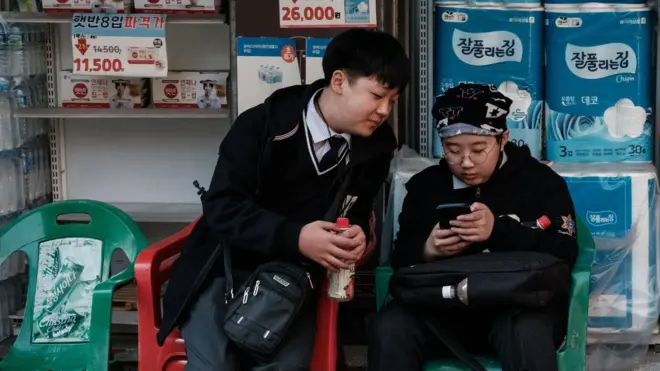 A South Korean schoolboy, wearing a school uniform and glasses, scrolls on his phone while sitting in a plastic chair outside a convenience store. Another boy in the same uniform leans over from his own chair to watch the screen.
