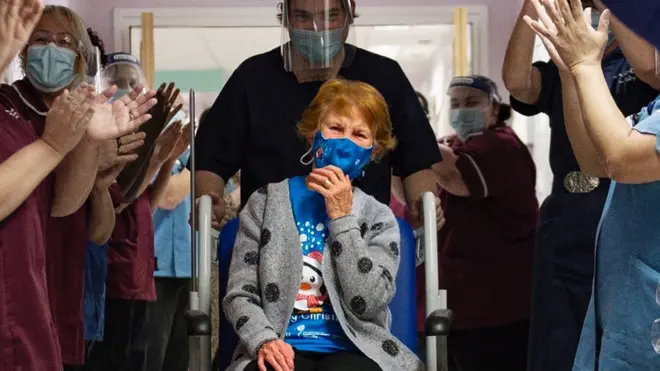 امرأة مسنة تضع كمامة جالسة على مرسي متنقل، فيما يصفق لها أشخاص في المستشفى.