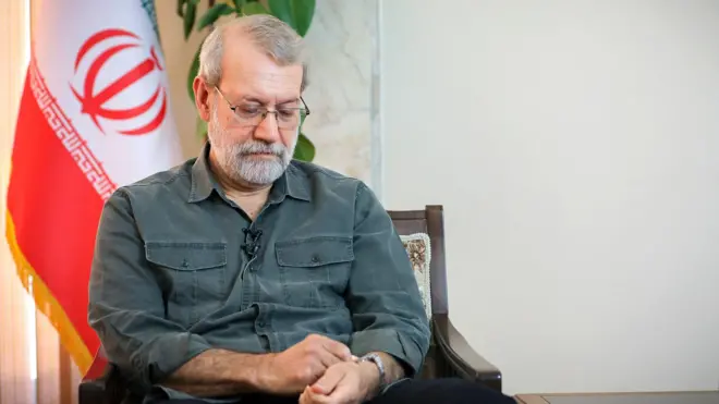 علی لاریجانی در مقابل پرچم ایران، در حال لمس و نگاه به ساعت مچی‌اش