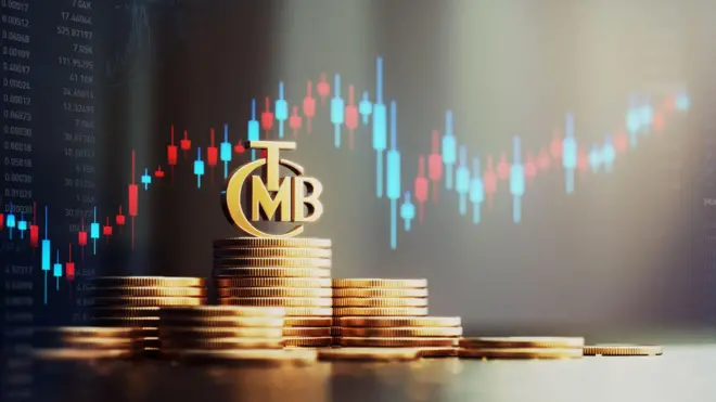 madeni paralar üst üste dizilmiş üzerinde TCMB amblemi var hepsi altın rengi ve arkada borsa işlemleri var mavi ve kırmızı bir grafik.