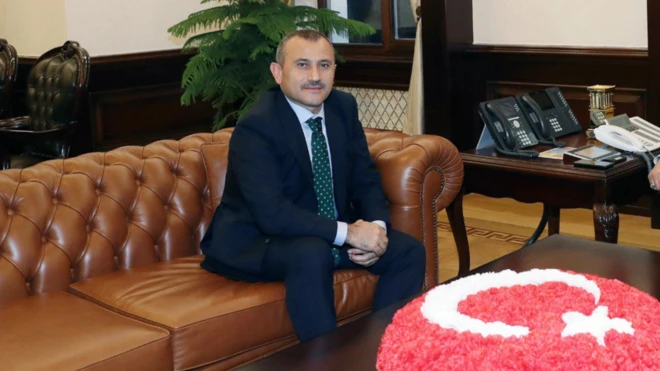 Kahverengi deri bir koltukta oturan bir takım elbiseli kişi görünüyor. Önünde Türk bayraklı bir koltuk var.