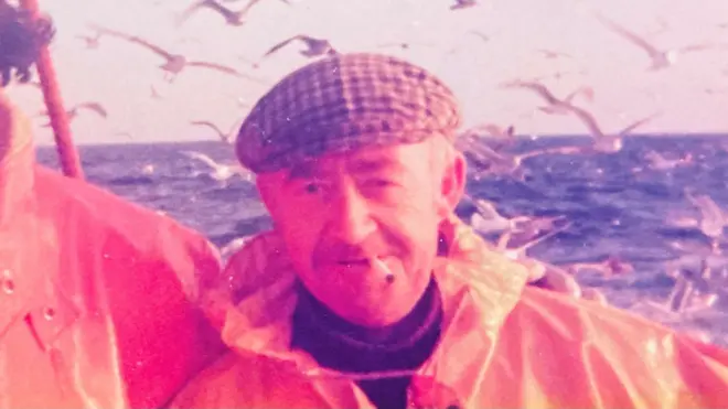 Old photo of fisherman on a fishing boat, wearing waterproof oils and a cap, the man - Jim Runcie - also has a cigarette in his mouth, sea and lots of seagulls in the background.