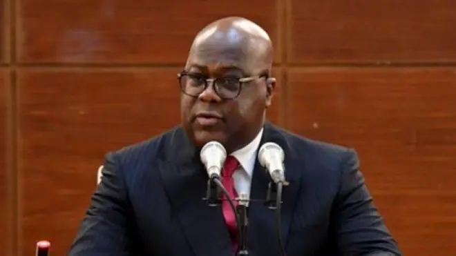 Perezida Félix Tshisekedi wa RDC arimo kuvuga ijambo yicaye imbere y'indangururamajwi ebyiri, inyuma ye hari urukuta rw'ibara ry'ikigina. Yambaye ikoti ry'umukara n'ishati y'umweru na karuvati ('cravate') y'umutuku ujya kuba iroza, n'indorerwamo. Ifoto yo ku wa gatandatu, i Kinshasa, ku itariki ya 18 Mutarama (1) mu mwaka wa 2025. 