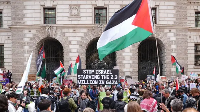A crowd of protesters standing and looking at the Foreign Office building, holding Palestinian flags and a sign that reads: Stop Israel starving the 2.3 million in Gaza