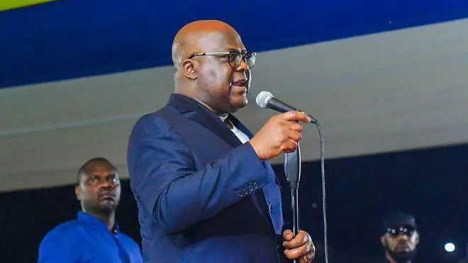 Perezida Félix Tshisekedi ahagaze imbere y'indangururamajwi arimo kuvuga ijambo, iruhande rwe hahagaze abagabo babiri, mu ruzinduko i Kisangani, ku wa gatatu, ku itariki ya 23 Ukwakira (10) mu mwaka wa 2024