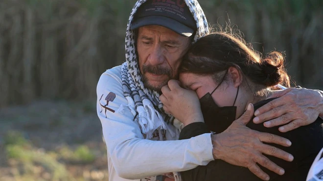 Женщина в черной санитарной маске плачет, уткнувшись в плечо мужчине, у которого на голове поверх кепки платок-арафатка