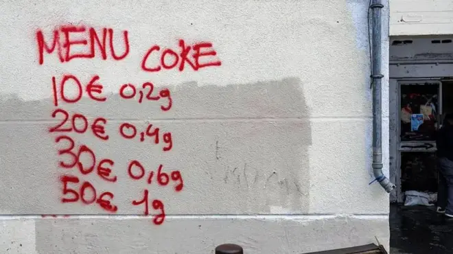 روی دیوار سفید نرخ یک ماده مخدر را با رنگ قرمز نوشته