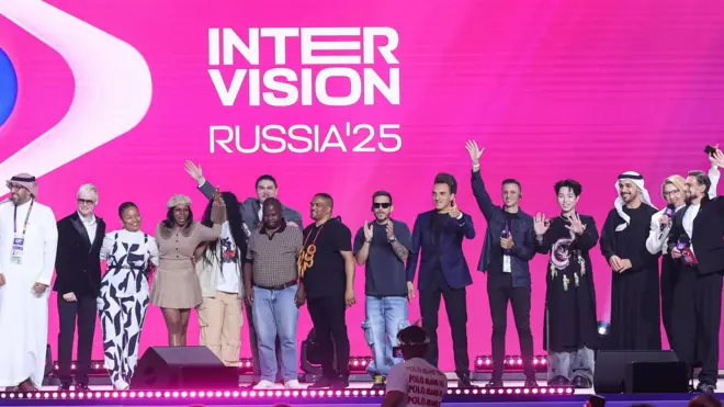 Os participantes do festival de música Intervision posam para as câmeras e acenam para o público de um palco. Atrás deles, um telão LED exibe o logotipo da Intervision Rússia 2025.