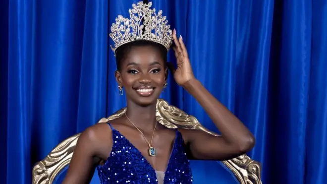 Miss Côte d'Ivoire 2022 Marlène-Kany Kouassi, qui porte une robe à paillettes bleu foncé avec des bretelles, touche sa couronne alors qu'elle est assise sur un trône avec un rideau bleu en peluche derrière elle.