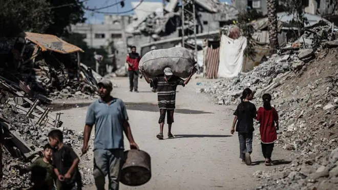 Pessoas andando em Gaza em meio a escombros. Uma delas carrega um saco nas costas 