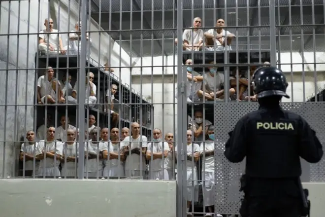 Presos com uniformes brancos estão na cela de uma prisão enquanto são observados por um policial 